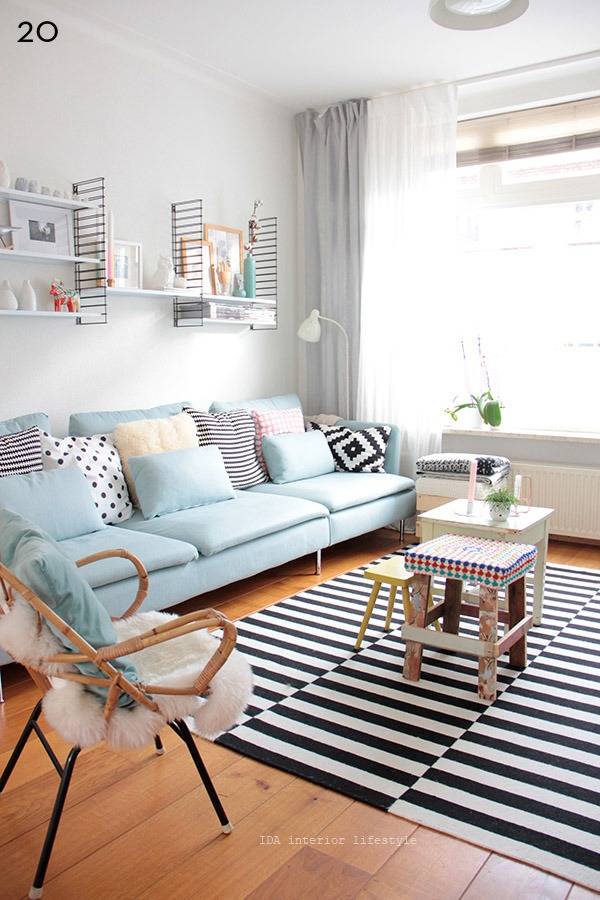 Pastel room arranged with blue color couch and wooden chair and tables rack shelves, potted plants and black and white striped carpets.