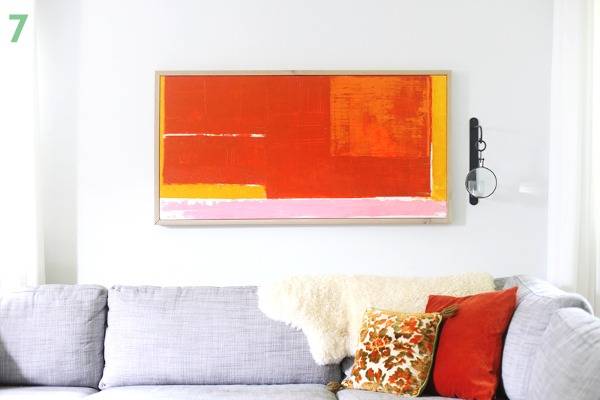 An orange painting is hanging over a white couch with pillows.