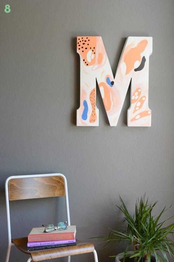 The letter M is hanging over a plant on a grey wall.