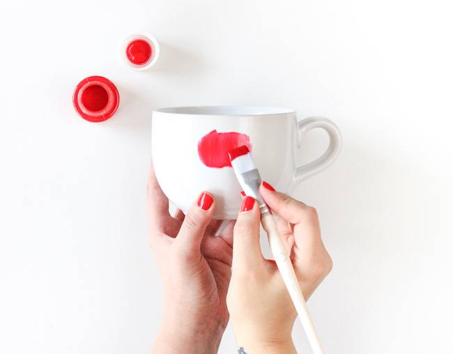 A person with red nails painting a dot on a white cup.