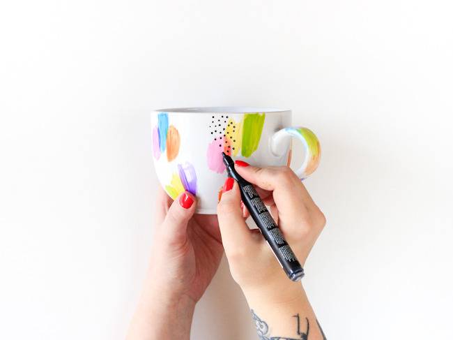 A person is using a marker to make a colorful design on a white cup.