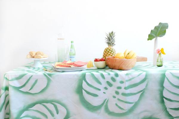 A giant leaf in green relief table cloth covering a table with a pineapple, a wooden bowl of yellow apples, a plate of meat sclices, a platter of bread rolls and a green glass bottle.