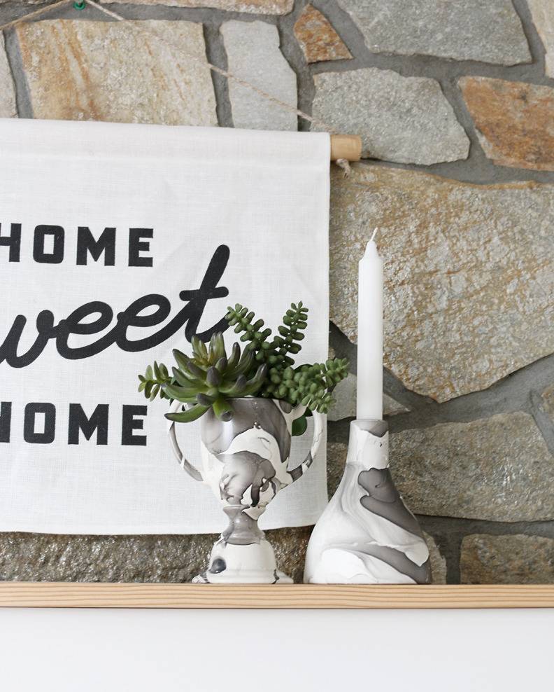 A home sweet home sign is above the mantle on a fireplace with a white candle in a candle holder next to it in a small plant.