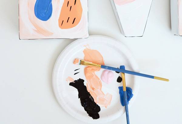 A paper plate with black, orange, pink and blue paint on it as well as two blue paintbrushes.