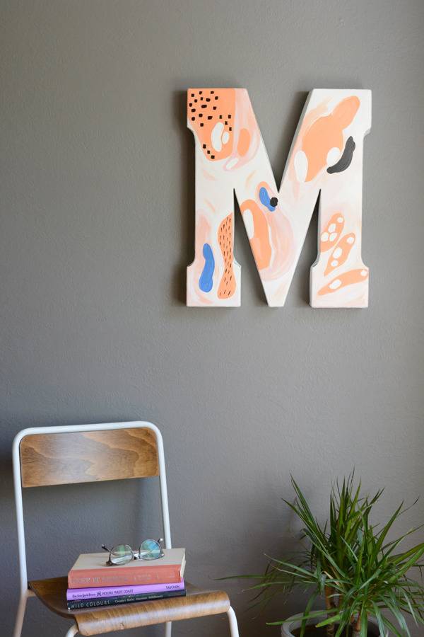 The letter M is hanging on a wall above a plant and a chair with books.