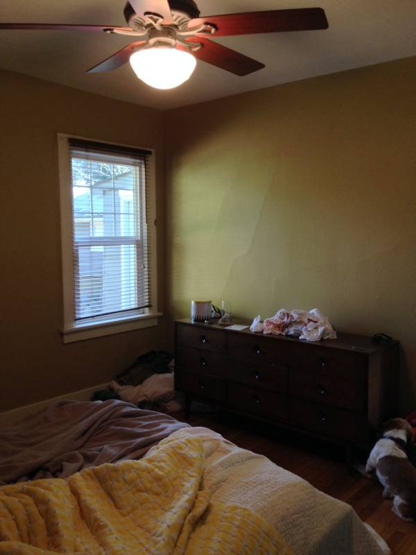 A messy olive green bedroom with a fan light turned on.