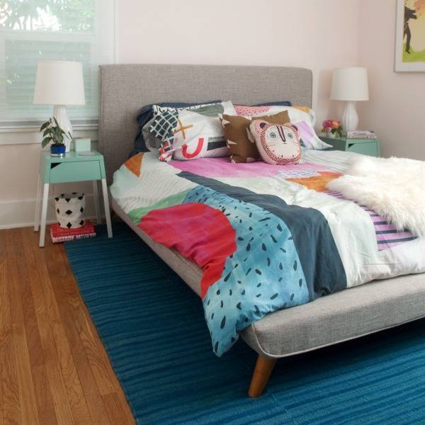 A bed with colorful sheets is sitting on a blue rug in a room with wooden flooring.