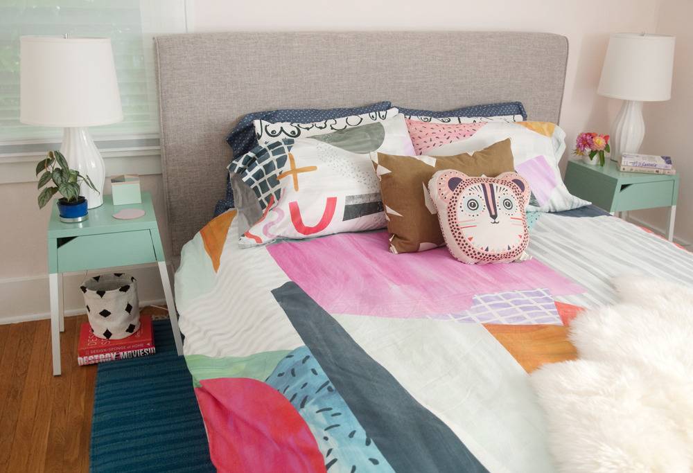 A bedroom with two light green side tables and matching white lamps that are on each side of acolorful pink, grey white, brown and red bedspread.