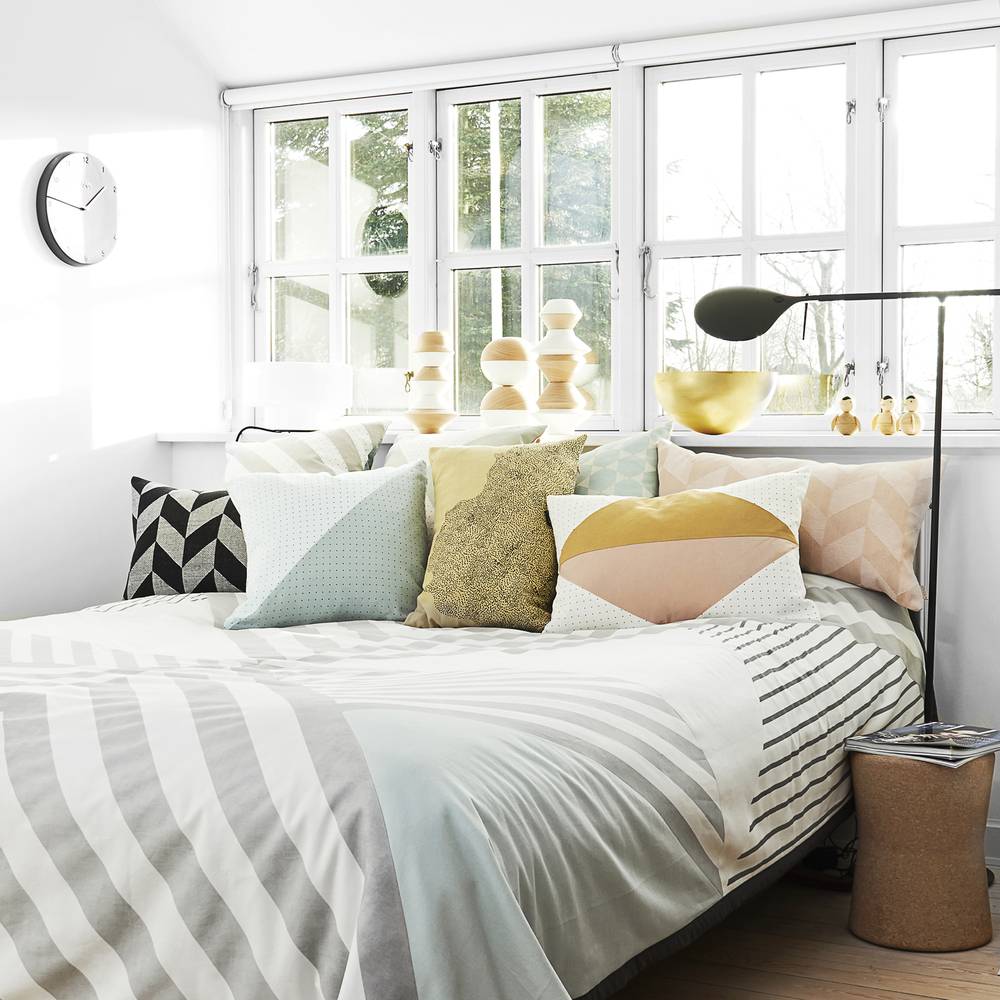 A bay of windows sit's behind a bed with a gray and white striped bedspread and colorful throw pillows.