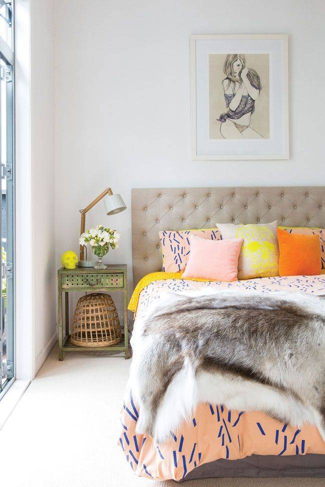 A bedroom with a pink coverlet covered by a grey fur blanket and pink, yellow and orange pillows on the bed.