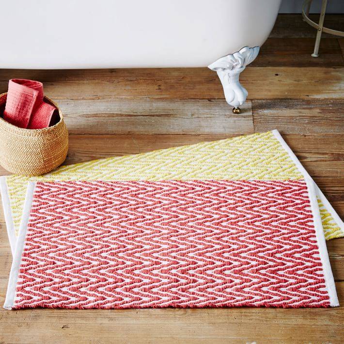 A woven red bath mat sits on top of a yellow one on a wooden floor and a bathroom near clawfoot tub.