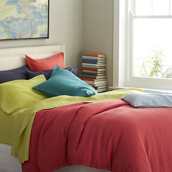 A colorful bedroom has a map of the bed that has red and yellow bedspreads purple, orange, and teal pillows, and a stack of colorful books is nearby.