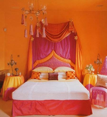 A middle eastern style bedroom has orange walls, and hot pink trim on the bed with the hot pink and orange tassel headboard of fabric.