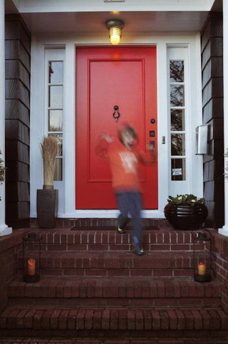 A boy in an orange shirt is coming down the stairs from a house with a red door.