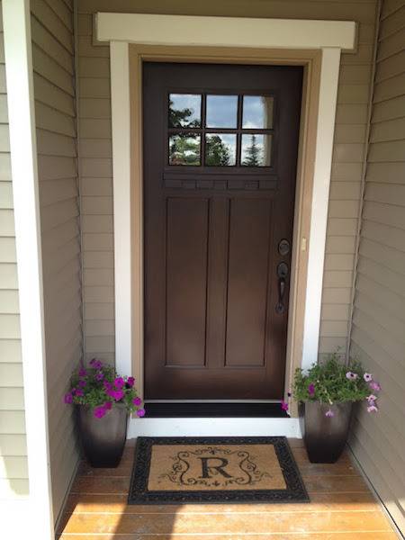 A doorway has two plants flanking the entrance and a welcome mat with the letter R on it.
