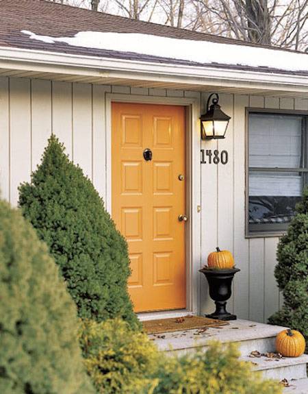The orange front door of a single story home with a pumpkin in a black vase on the landing.