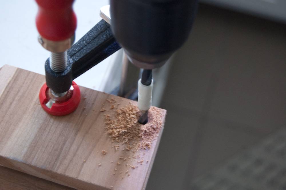 A block of wood clamped with a red clamp is being drilled in its corner.