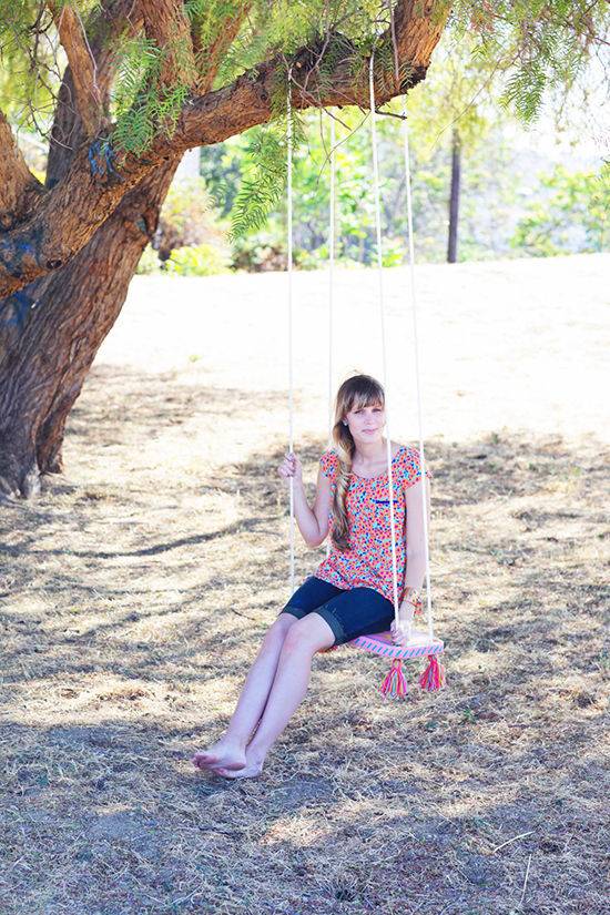 A young lady in pink and blue sits on a chair swing under a large shady tree.