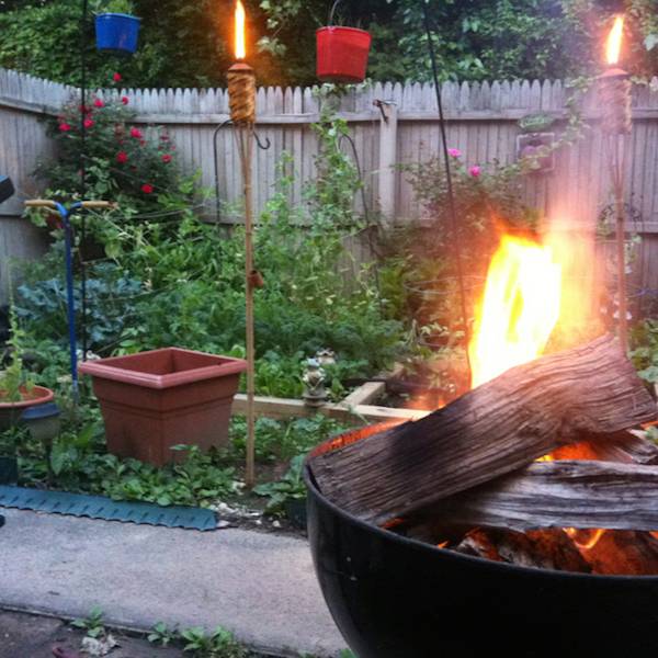 A fire glows in a firepit in a garden area outside near a fence.