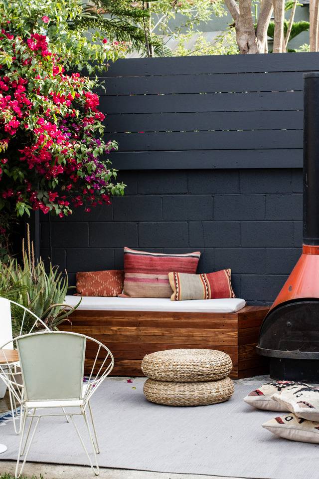 An ottoman sits in front of a wooden bench with throw pillows on it in front of a black curtain on a patio.