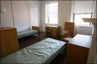 There are two empty beds along with desks and chairs in the dorm room.