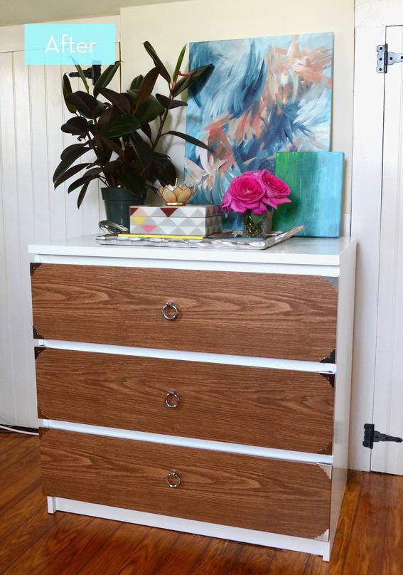 A brown and white three drawer dresser with a black plant, pink flowers and a blue abstract painting on top of it.