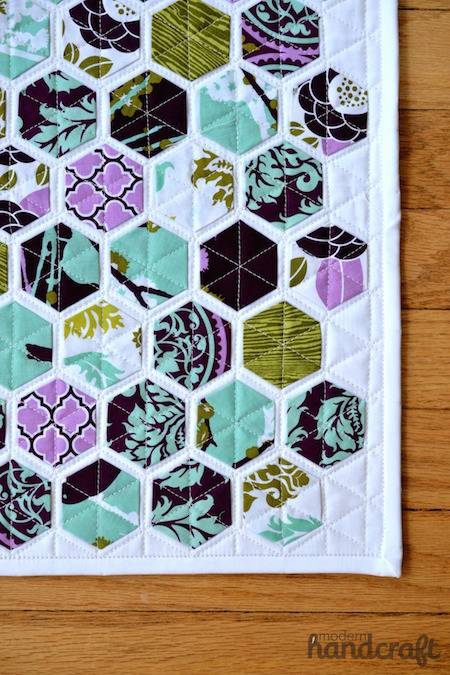 Modern hexagon quilt with purple and light blue colors sitting on top of a wooden floor.