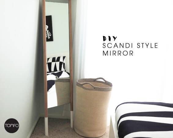 Scandi style mirror resting up against a wall in front of a small clothes hamper in a bedroom.