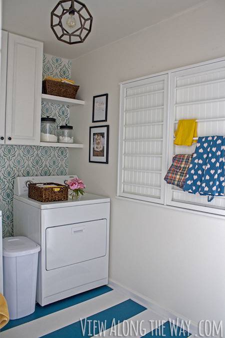 A white laundry room has a washer, trash and shelves.