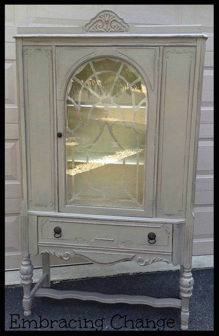 A white antique dining room hutch with a window on the front sits in front of a white brick wall.