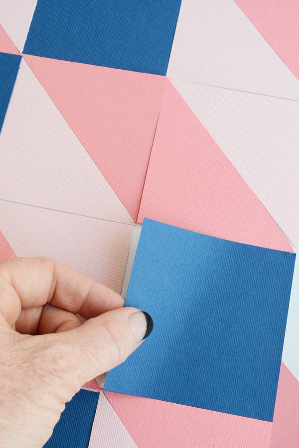 A hand hold a square blue piece of paper above pink and white pieces of square paper.