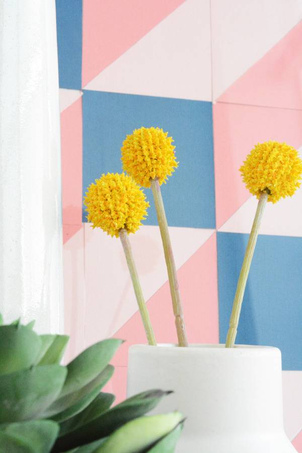 A flower with yellow balls is sitting in a cup near a colorful pastel wall.