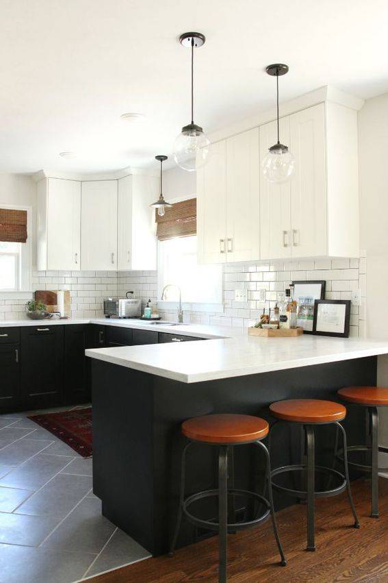 A pristine white kitchen with a white counter and three brown topped round stools under its lip and with two silver pendulant lights with large clear round bulbs hanging over it.