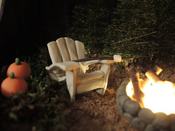 A small off-white chair sits near a campfire and has two pumpkins off to the side.