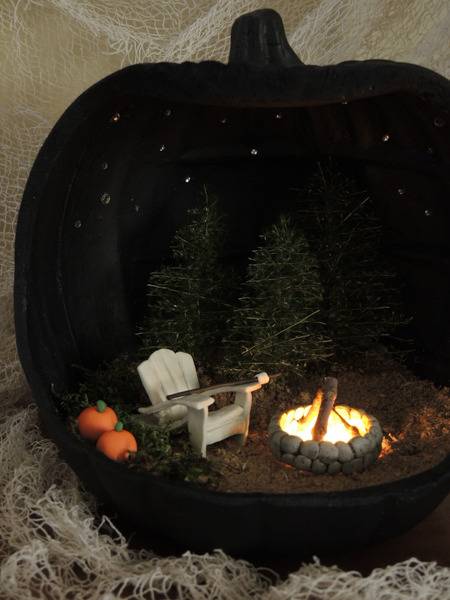A stone fire pit with a fire in it, next to a white adirondak chair and two small pine trees, with two pumpkins next to the chair and all inside of a black pumpkin.