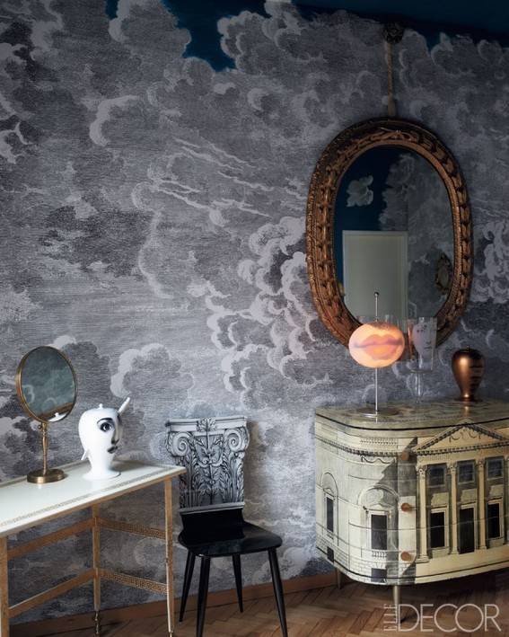 A table and black chair sit in a corner of a room with grey cloudy walls and an old counter and mirror.