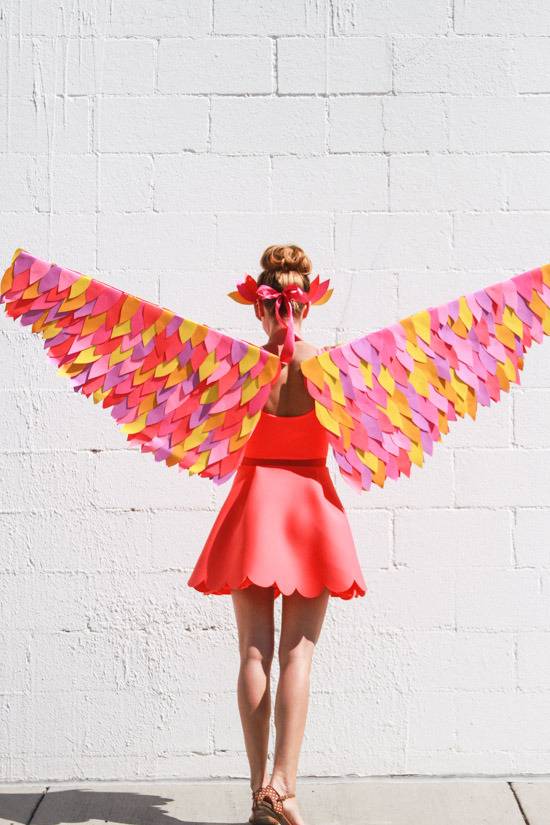 The back of a woman with orange,pink, purple and yellow wings spread and an orange skirt standing in front of a white stone wall.