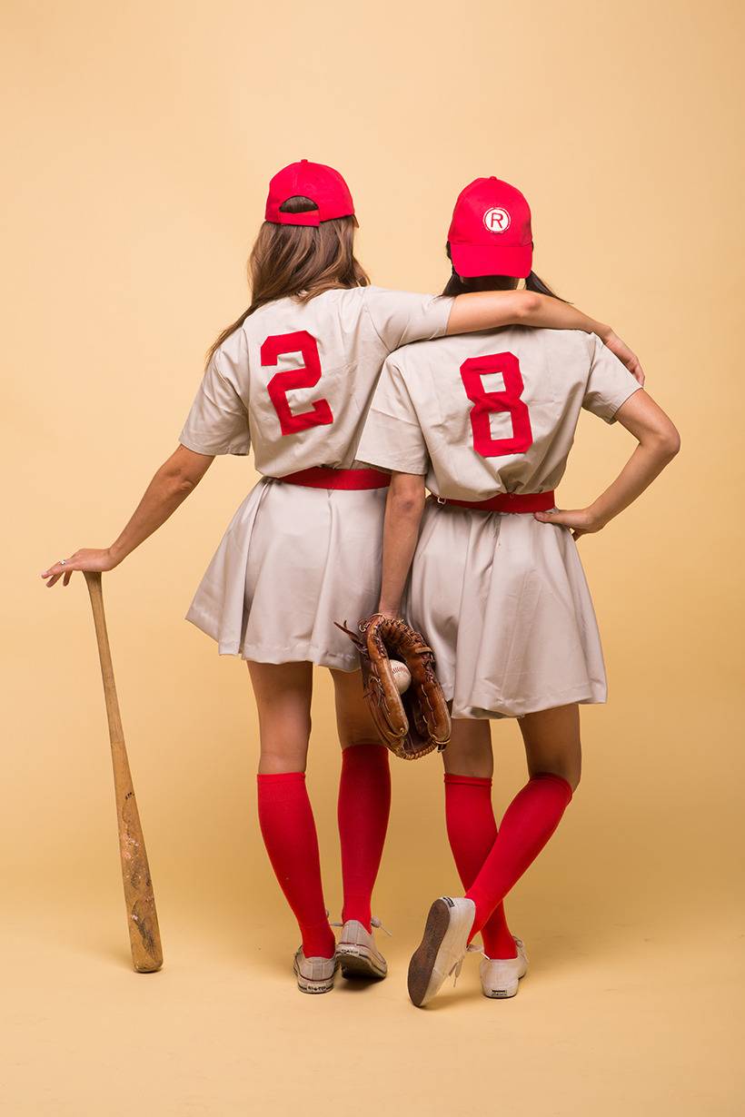 Two girls dressed in sports uniform are standing and one of them is holding a baseball bat.