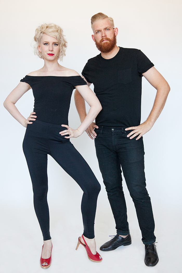 A couple wearing all black stand and pose next to each other.