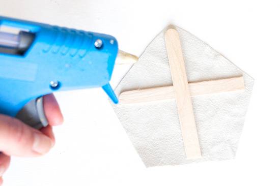 A woman's hand holding a blue glue gun, poised to glue popsicle sticks onto white paper.