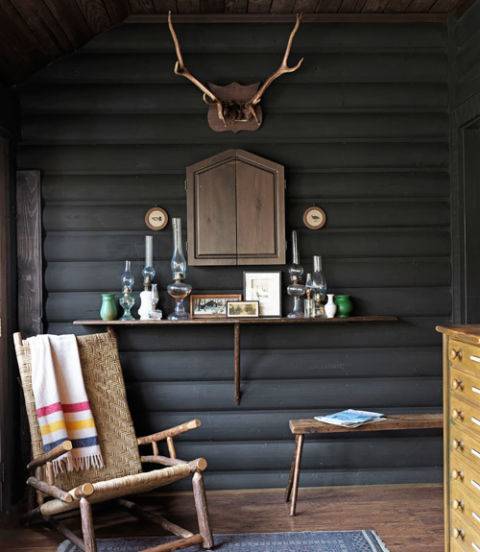 A wicher rockig chair in front of a grey log wall with a pair of antlers hanging on it above a shelf of candles.