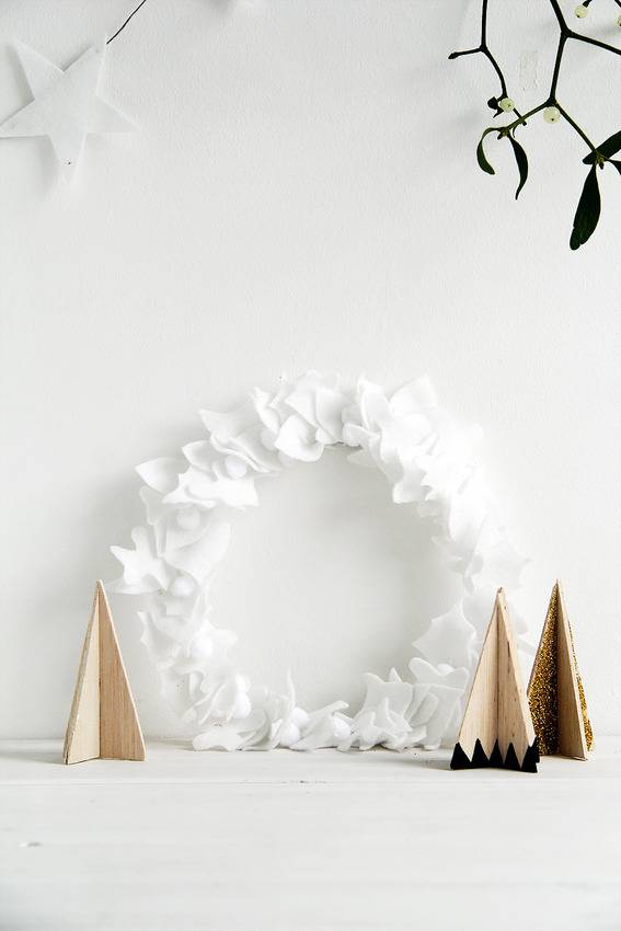 A white wreath against a white wall with a white star on the wall and three gold craft trees in front of the wreath.