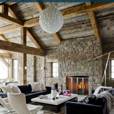 The interior of a stone cabin with with a high wooden ceiling and a large white globe pendant light, a fireplace and two black sofas.