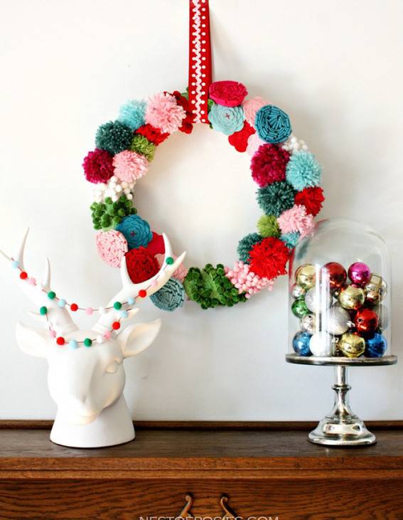 A two ring multicolored wreath hangin on a white wall behind a white reindeer head statue with colorful antlers and a covered cake plater filled with Christmas balls.