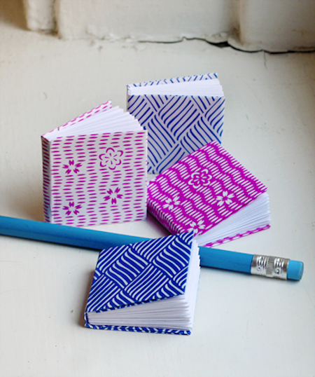 Miniature books sit on a surface near a blue pencil.
