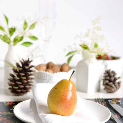 A pear on a white folded napkin on a white plate in front of a pine cone on another plate with a white vase and green leaves.