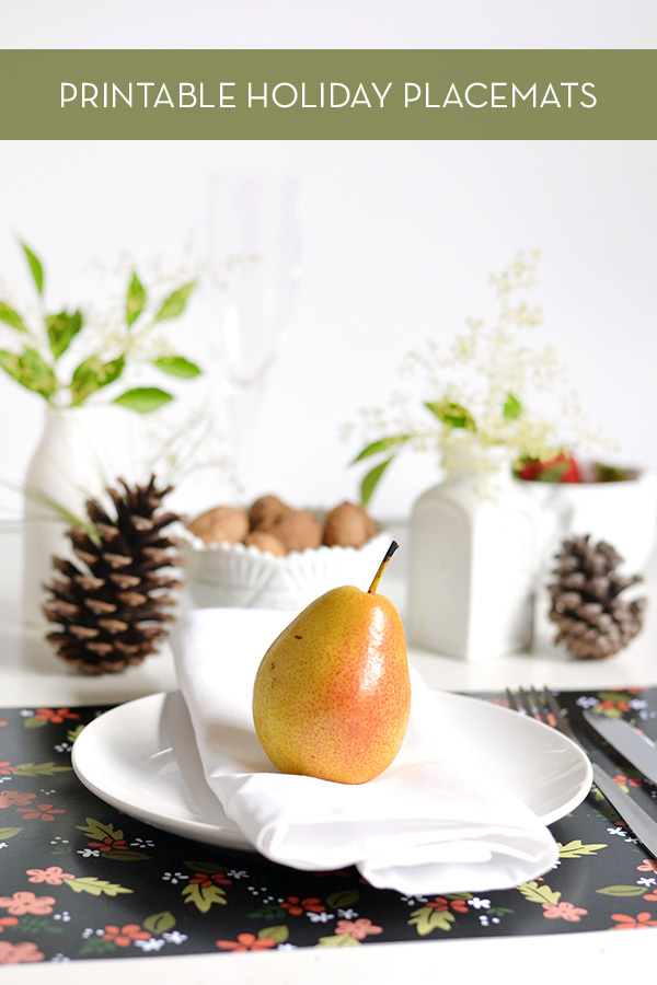 A pear on a white folded napkin on a white plate in front of a pine cone on another plate with a white vase and green leaves.