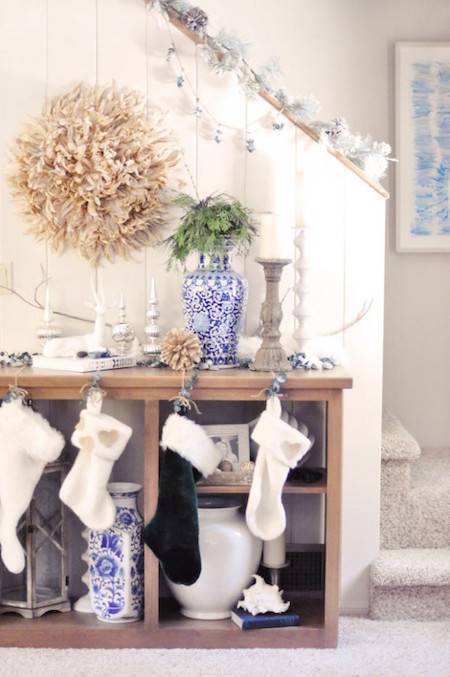Christmas stockings hanging on a wooden bookshelf decorated with a vase, wreath and candle stick, next to a staircase in a home with white walls and white carpeting.
