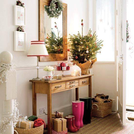 There are boots lined neatly beneath a side table that holds a plant and decor beneath a mirror where a wreath is displayed.