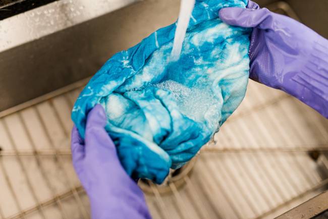 Person holding a tie dyed cloth under a running faucet while wearing purple rubber gloves.